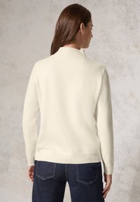 Maglione color crema con colletto alto, polsi e orlo a coste, realizzato in un materiale a maglia testurizzato, abbinato a jeans in denim scuro.