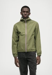 Veste imperméable vert olive avec capuche, poignets élastiques et fermeture éclair sur le devant avec des accents multicolores. Associée à un jean foncé.