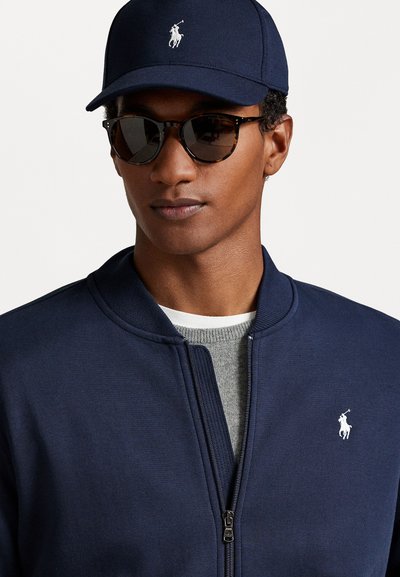 Jūras flotes zip-up jaka ar ribotu apkakli, ar baltu logotipu. Pelēks džemperis zem tās un melnas saulesbrilles ar bruņurupuču apmali.