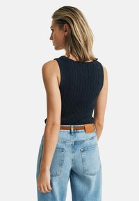 Marineblauwe kabelgebreide mouwloze top, gecombineerd met lichtblauwe high-waisted jeans, voorzien van een tan leren riem met reliëflogo en achterzakken.