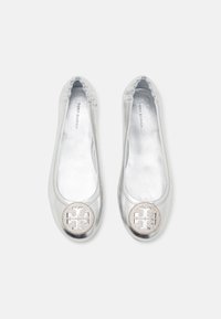 Bailarinas plateadas de Tory Burch con bordes elasticados y medallones con logo redondo en las puntas, exhibidas lado a lado sobre un fondo blanco.