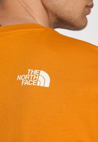 Tricou din bumbac portocalie cu mâneci scurte, prezentând un logo alb "The North Face" pe partea superioară a spatelui. Textură netedă, croi regular.