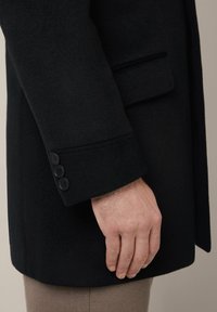 Czarny wełniany płaszcz o dopasowanym kroju; posiada pojedynczy mankiet z trzema czarnymi guzikami oraz subtelną teksturą. Rękaw sięga do połowy nadgarstka.