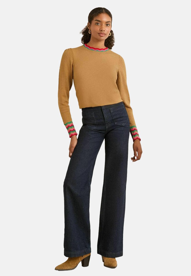 Beige trui met geribbelde lichtrode, groene en roze golfranden, gecombineerd met donkerblauwe wijde jeans en beige hoge enkellaarsjes.