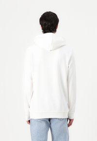 Sweat à capuche blanc avec une coupe décontractée, des poignets et un ourlet côtelés, associé à un jean bleu clair. Texture lisse et détails de design minimaux.