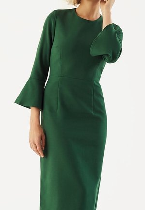 Donna che indossa un vestito aderente verde scuro a maniche lunghe con polsini svasati e scollo rotondo, su sfondo semplice.