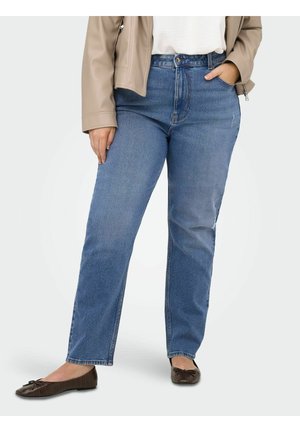 Person iført blå cowboybukser med høj talje, beige jakke, hvid top og mørkebrune flade sko, stående med den ene hånd i lommen.