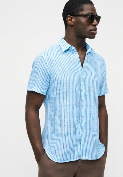 Man wearing tortoiseshell sunglasses and a light blue short-sleeve shirt with thin vertical blue stripes, standing with hands in pockets.