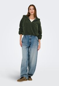 Sudadera con capucha de forro polar en verde oscuro, con cuello con cremallera, combinada con jeans de pierna ancha en azul claro y zapatos de leopardo de slip-on.
