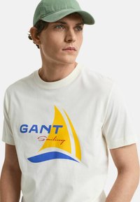 Witte katoenen t-shirt met een blauw en geel zeilbootafbeelding en de tekst "GANT Sailing" in het rood. Model draagt een groene pet.