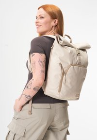Beige Roll-Top-Rucksack aus robustem Material, mit einer Vordertasche mit Reißverschluss und verstellbaren Trägern. Glatte Textur mit subtilen Akzenten.