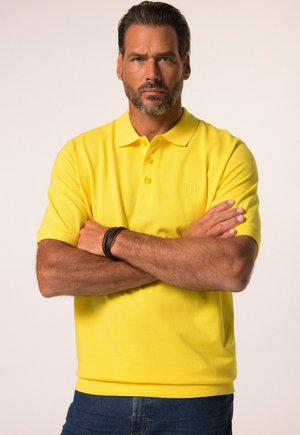 Polo gialla con colletto, dotata di tre bottoni e un sottile dettaglio con logo. Il tessuto sembra liscio, abbinato a jeans blu.
