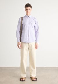 Lichtpaarse overhemd met knopen, crèmekleurige broek en bruine sandalen met zwarte zolen. Accessoires omvatten een bruine schouderband.