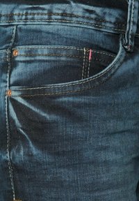 Jeans de mezclilla oscura con un bolsillo trasero con remaches de cobre y costuras en contraste en color rosa. Textura suave con un leve desgastado.