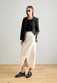 Blazer nero su misura, top nero aderente, gonna midi beige a portafoglio con cucitura diagonale e scarpe in pelle lucida nere. Occhiali da sole rotondi.