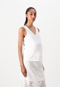 Weißes Spitzentop für Schwangere mit V-Ausschnitt und ärmellosem Design, über einem passenden Spitzenrock mit floralem Muster und strukturiertem Stoff.