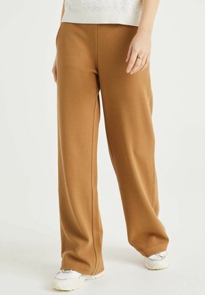 Persoon in gebruinde, gestructureerde, wijde broek, witte sneakers en een witte top, met ontspannen handen langs de zijden.