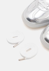 Baskets argentées pour tout-petit avec des lacets blancs enroulés à côté, sur un fond blanc.