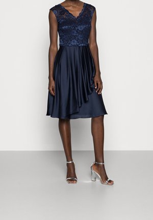 Vrouw draagt een mouwloze marineblauwe kanten topjurk met een asymmetrische satijnen rok, gecombineerd met zilveren sandalen met hoge hakken, staand op een effen achtergrond.