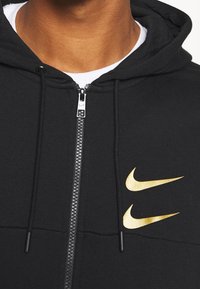 Black zip-up hoodie with gold Nike logo accents, featuring a hood, drawstrings, and textured fabric detail along the zipper.