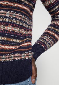 Stickad tröja med en marinblå bas och flerfärgade mönster i rött, vitt och grönt. Ribbad mudd och nederkant, mjuk textur.