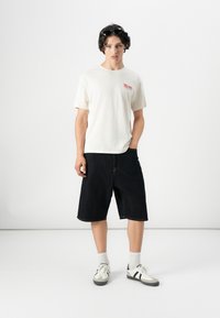 White cotton t-shirt with a small red graphic, paired with knee-length dark denim shorts and white sneakers with black accents.