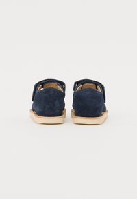 Zapatos de ante azul marino con una correa de velcro, parte trasera redondeada y una suela de goma de color marrón claro. Textura suave con detalles de costura mínimos.