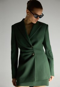 Vihreä muotoiltu bleiseri, jossa on kapea vyötärö, kaksinkertaiset napit ja rakenteelliset hartiat. Materiaali näyttää sileältä ja teksturoidulta.