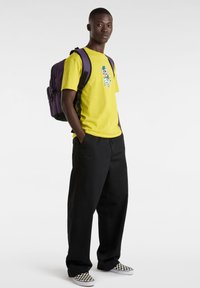 Camiseta amarilla con un diseño gráfico, pantalones anchos negros y zapatos slip-on a rayas en blanco y negro. Mochila morada con correas ajustables.