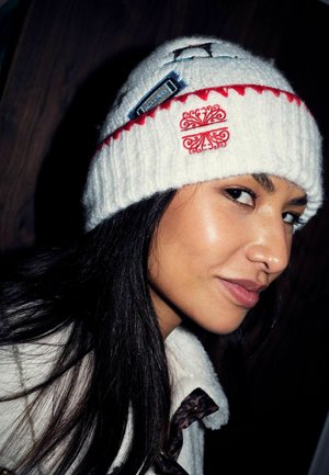 Proua pikkade tumedate juustega naine, kandes valget kootud mütsi punase tikandiga ja valget fliisjakki leopardimustriga detailidega.