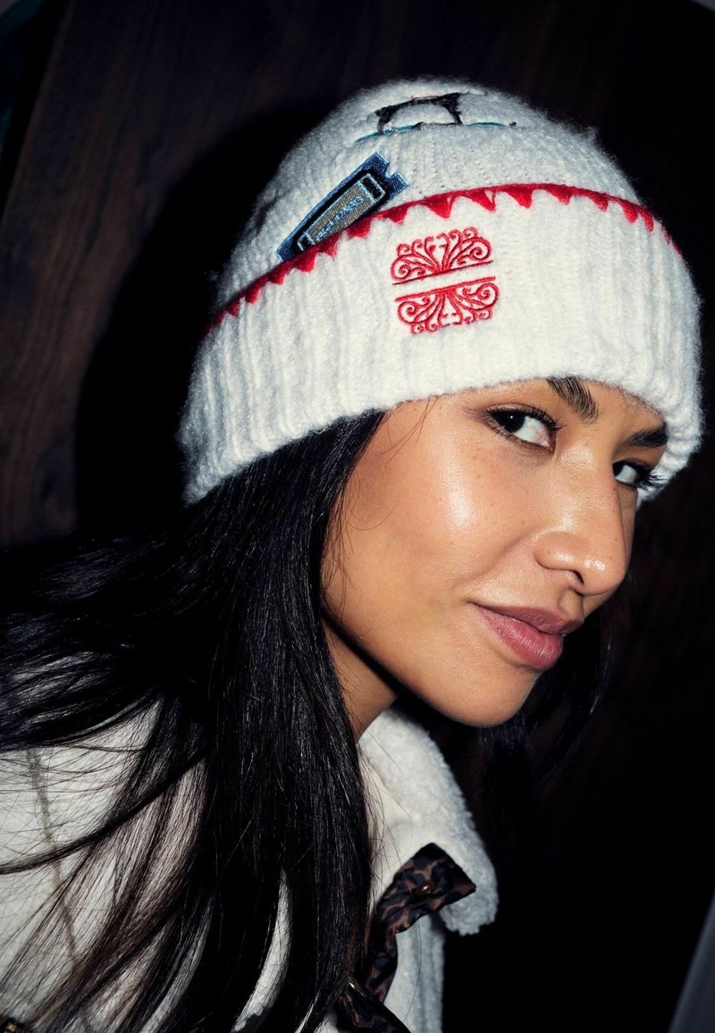 Femme aux longs cheveux bruns portant un bonnet en tricot blanc avec une broderie rouge et une veste polaire blanche avec un motif léopard.