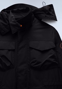 Black jacket with high collar, two front flap pockets, and detachable hood with button closure, laid flat on white surface.