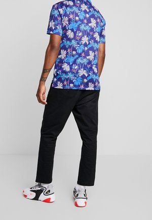 Homme debout, dos à nous, portant une chemise bleue imprimée tropicale, un pantalon noir et des baskets blanches avec des touches rouges sur fond gris.