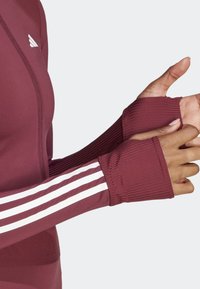 Närbild av en person som bär en vinröd långärmad träningströja med vita ränder och tumhål på ärmarna.