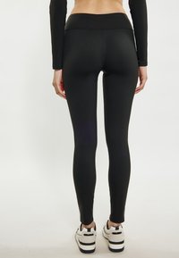 Schwarze Leggings mit hohem Bund, aus einem glatten, dehnbaren Material. Das Model trägt weiße und schwarze Turnschuhe.