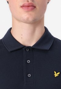 Tumši zils polo krekls ar teksturētu audumu, trīs priekšējām pogām un dzeltenu izšūtu ērgļa logotipu kreisajā krūšu pusē.