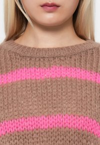 Brązowy sweter dzianinowy z teksturowanym wzorem i dwoma poziomymi jasnoróżowymi paskami na klatce piersiowej. Okrągły dekolt.
