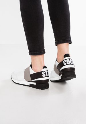 Baskets slip-on blanches et grises avec un design texturé, des accents noirs en contraste et un texte imprimé audacieux à l'arrière. Semelle basse.