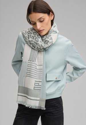 Femme portant une veste bleu clair et une écharpe à motifs géométriques et imprimé léopard, debout devant un fond gris uni.