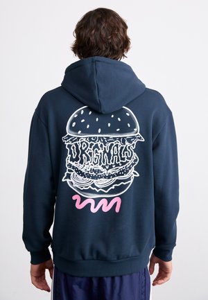 Personne portant un sweat à capuche bleu marine avec un graphisme de hamburger blanc et le mot "ORIGINALS" au dos, debout devant un fond blanc uni.