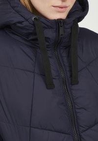 Jachetă puffer bleumarin cu închidere cu fermoar, având un model matlasat, șnururi negre și un guler înalt pentru o căldură suplimentară.