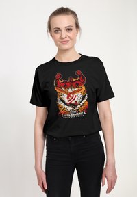 Žena nosící černé tričko Captain America Brave New World s třemi ilustracemi superhrdinů v červené, bílé a oranžové barvě.