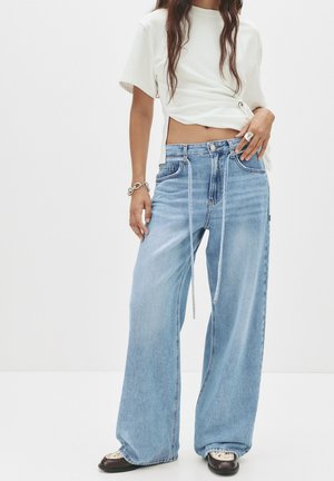 Jean large bleu clair à taille haute, avec poches avant et détail à cordon. Assorti avec un t-shirt blanc court et des chaussures noires.