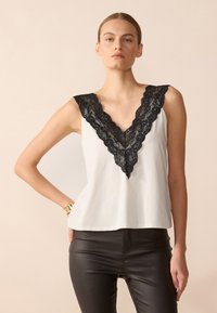 Top sin mangas blanco con un cuello en V de encaje negro, tela suave y ajuste relajado. Combinado con pantalones negros ajustados.