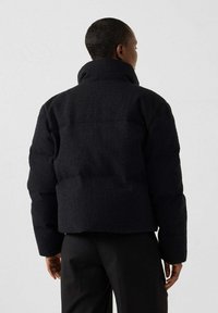 Personne portant une veste noire texturée à col montant et un pantalon noir, de dos contre un fond gris clair uni.