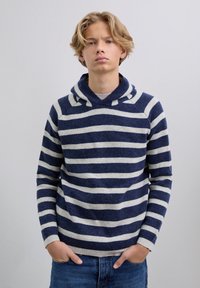 Nuori, vaaleahiuksinen henkilö, joka on pukeutunut laivastonsiniseen ja valkoiseen raidoitettuun huppariin, seisoo kädet sinisten farkkujen taskuissa yksinkertaista taustaa vasten.