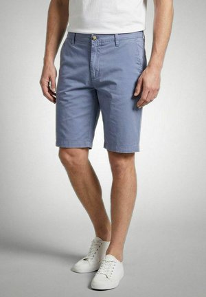 Man wearing light blue knee-length shorts and white sneakers, standing against a plain grey background.