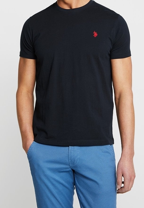 Svart bomull T-shirt med rund hals och korta ärmar, med en liten röd logotyp på vänster bröst. Bärs med blå texturerade byxor.