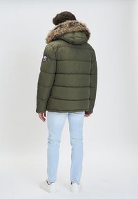 Olivenfarvet pufferjakke med en pelsforet hætte, med kviltede mønstre. Båret over lyseblå jeans og hvide sneakers, set fra ryggen.