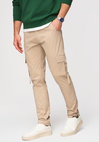 Homme portant un pantalon cargo beige, des baskets blanches, un sweat vert et une montre avec un cadran bleu, debout, la main gauche dans la poche.